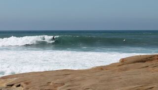 Waves breaking while surfers paddle out, seen from a cliff